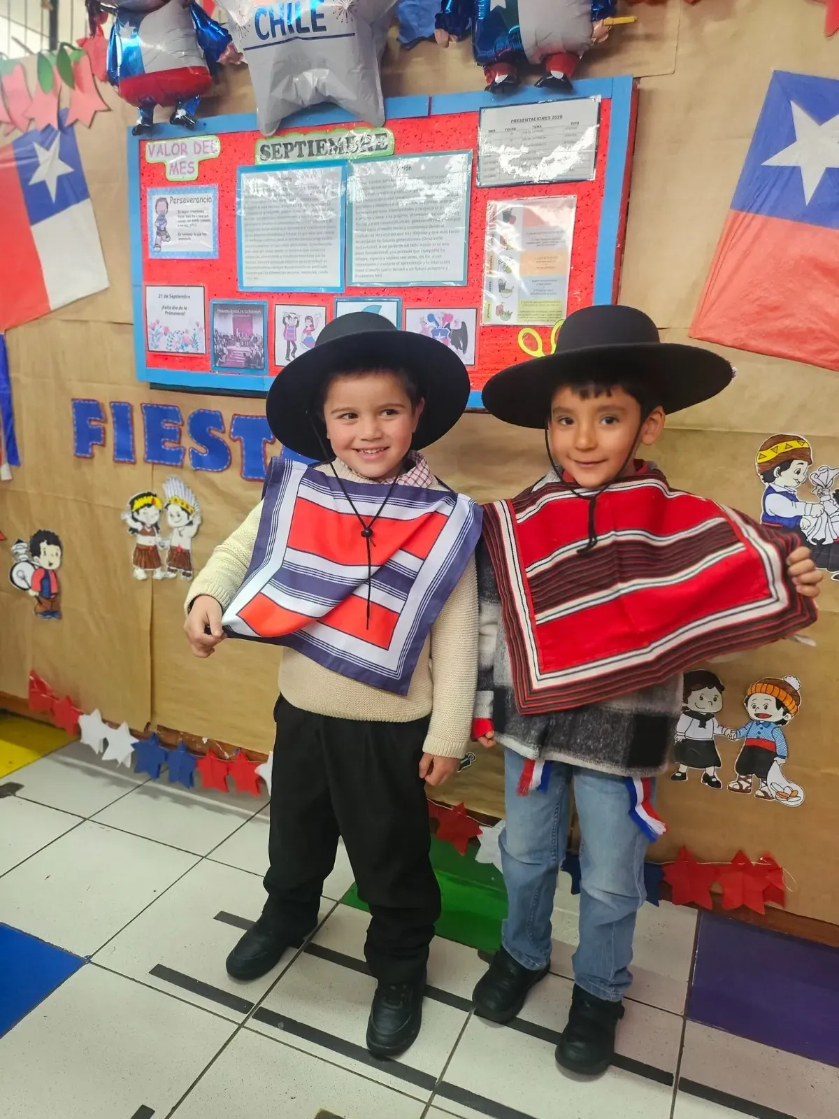 Dos párvulos sonrientes vestidos de huasitos posando frente al diario mural de Septiembre en sala