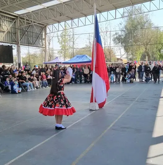 Párvulo con vestido de china y pañuelo bailando cueca junto a la bandera chilena